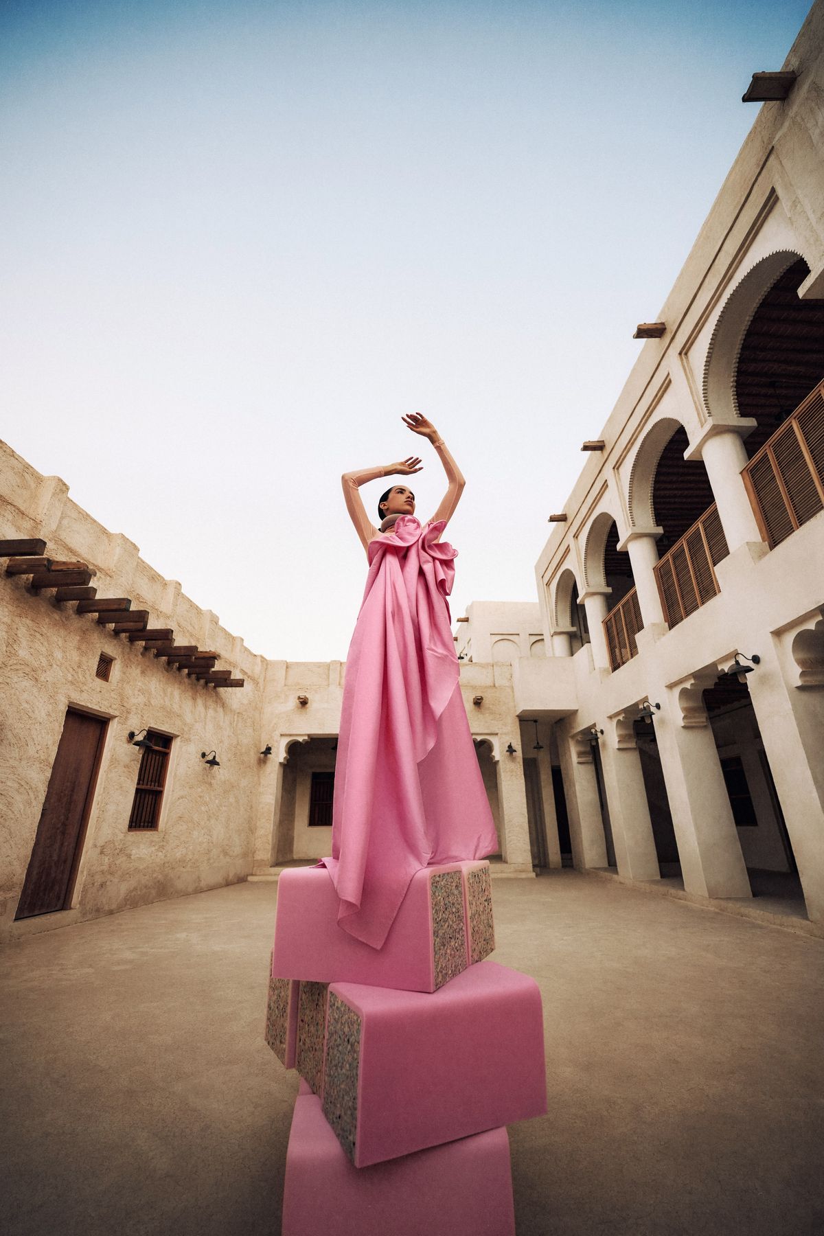 Model in a pink gown standing atop stacked blocks in a Qatari courtyard