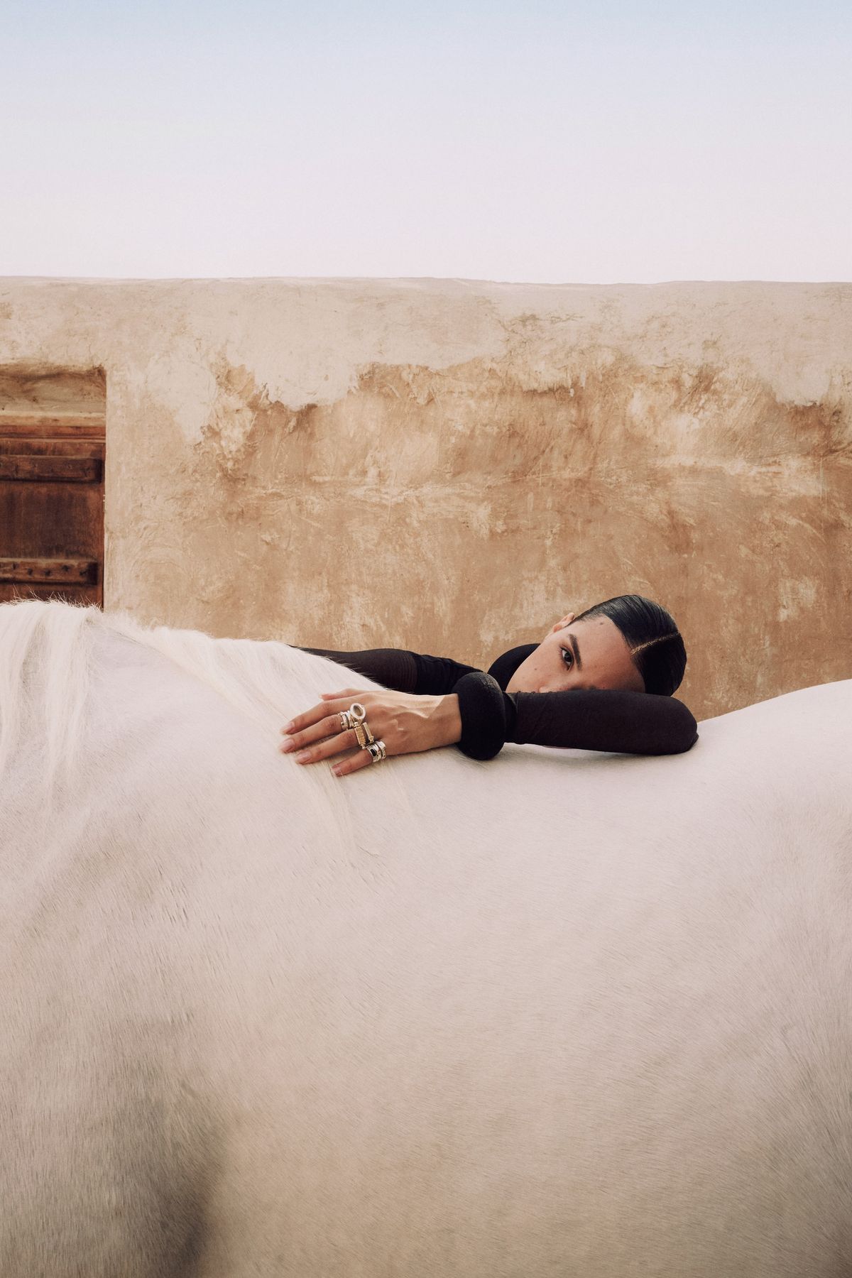 Model resting against the back of a white horse beside a sand-coloured wall